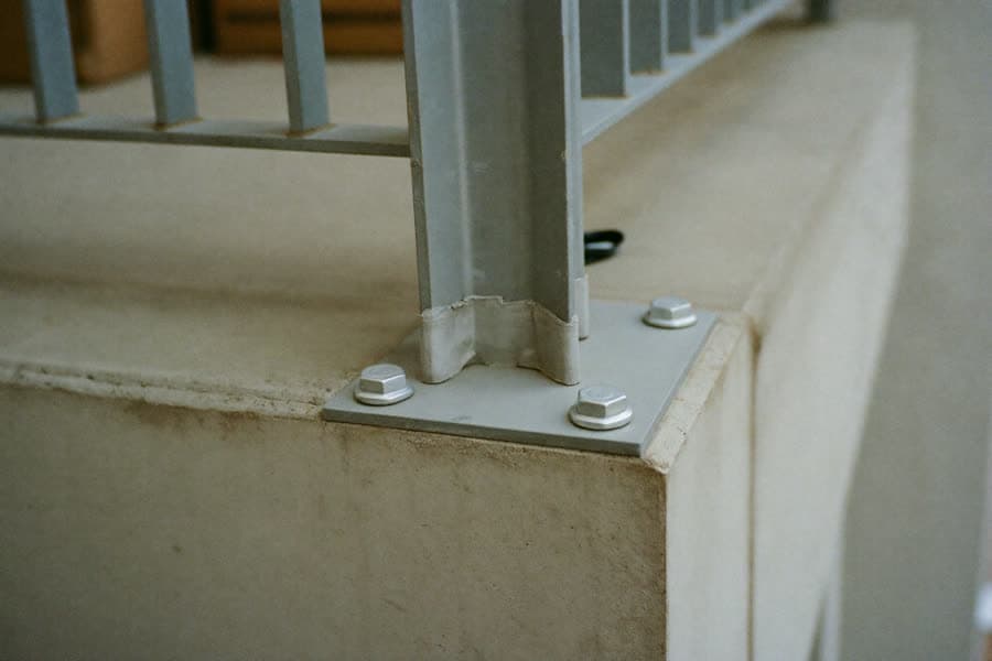 Befestigungspunkt eines Balkongeländers: die Fußplatte eines Pfostens ist mit vier Sechskantschrauben auf der Betonbrüstung verankert, um den Pfostenfuß liegt eine Dichtmanschette
