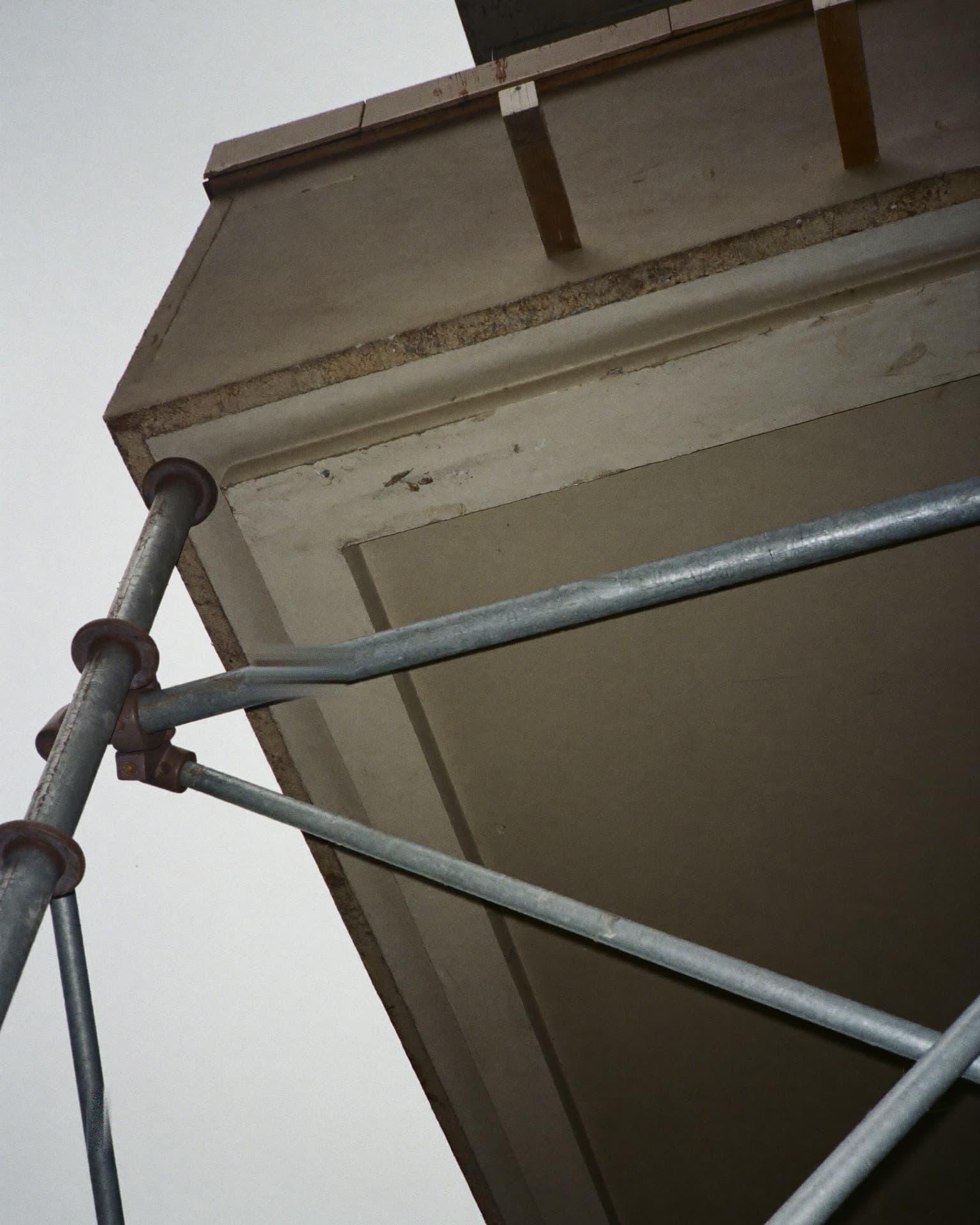 Blick von unten unter eine Betonbalkonplatte: die graue Untersicht und die Plattenstirn laufen schräg durch das Bild, davor kreuzen verzinkte Gerüstrohre mit Kupplungen, oben liegen Bohlen auf, dahinter heller, bedeckter Himmel