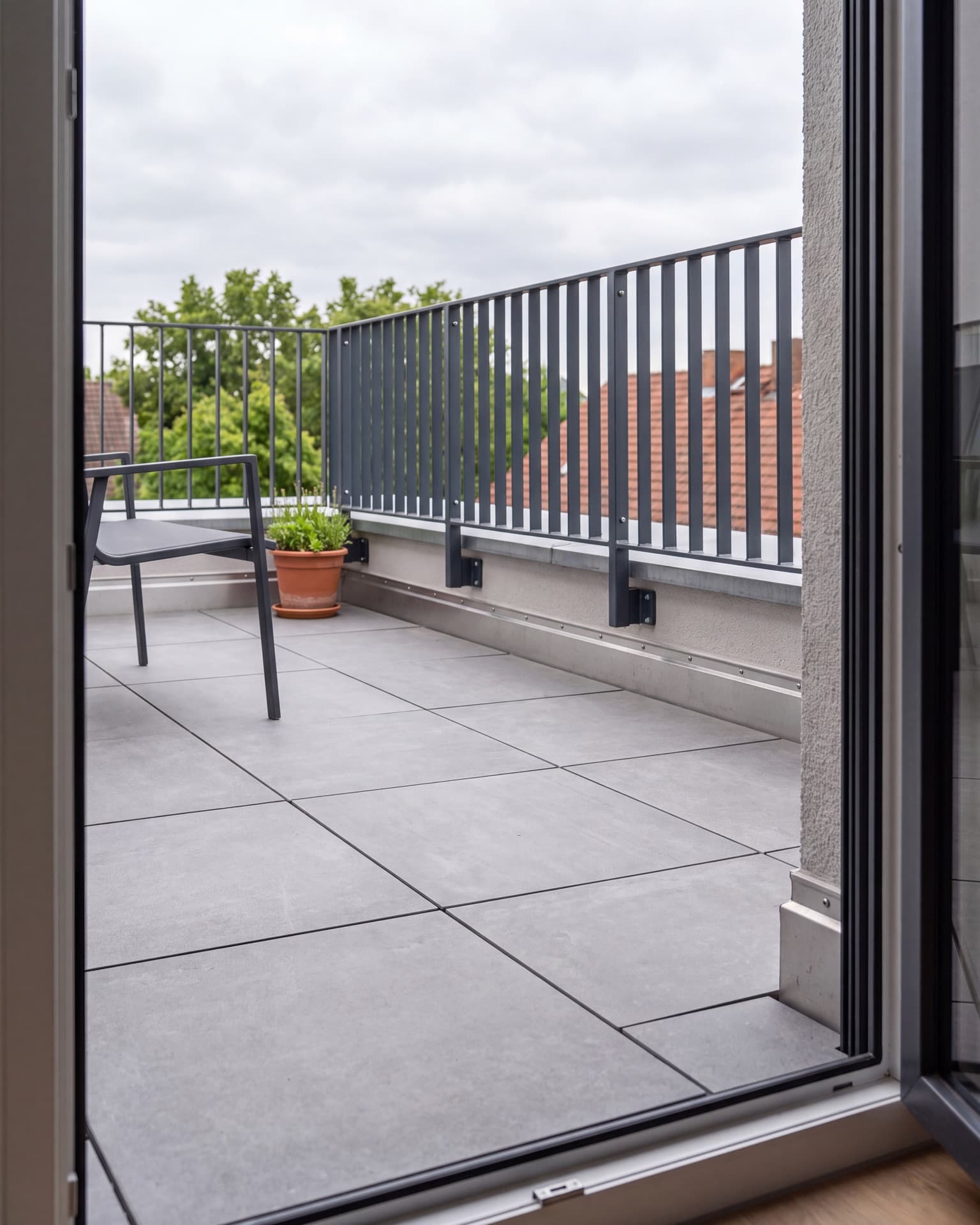 Blick von der offenen Wohnungstür auf einen fertig sanierten Balkon: großformatige graue Platten auf Stelzlagern mit offenen Fugen, an der Wand eine hochgeführte Abdichtung hinter schmaler Metallkante, am vorderen Rand ein anthrazitfarbenes Stabgeländer, dahinter Baumkronen und Nachbardächer; seitlich ein Stuhl und ein bepflanzter Topf, bedeckter Himmel